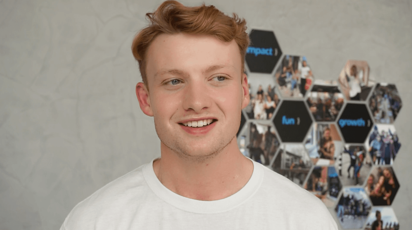 Een jonge man die werkt als klantadviseur bij Independer kijkt glimlachend naar rechts en staat voor een muur met foto's van Work-on, aangezien hij werkzaam is via Work-on bij Independer.