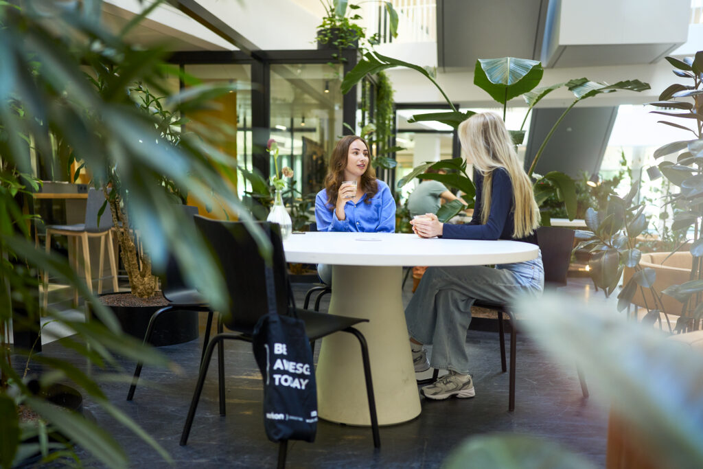 Twee vrouwen zitten aan een ronde tafel in een lichte, moderne ruimte met veel groene planten. De vrouw links draagt een blauwe blouse en houdt een glas vast, terwijl de vrouw rechts een donkerblauwe top en wijde jeans draagt. Er hangt een tasje aan een van de stoelen met de tekst 'Be awesome today'.