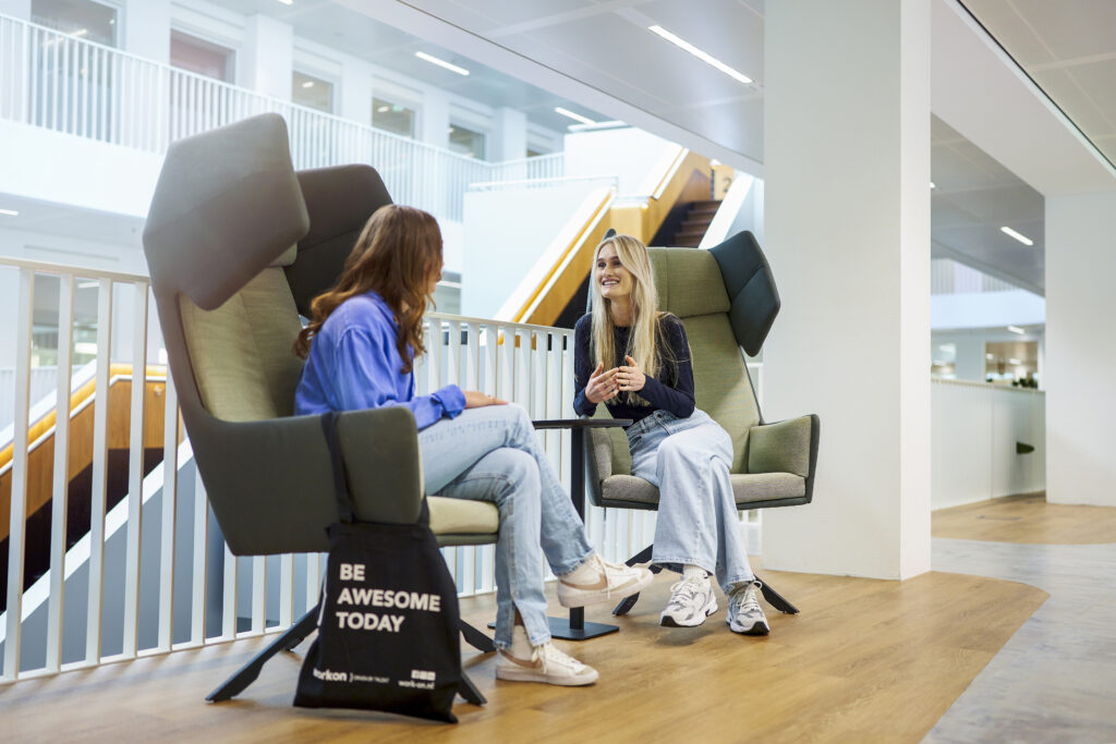 Twee jonge vrouwen zitten kletsend in comfortabele stoelen, in een lichte, open kantoorruimte. Er staat een tas van Work-on op de grond met de tekst 'Be awesome today'.