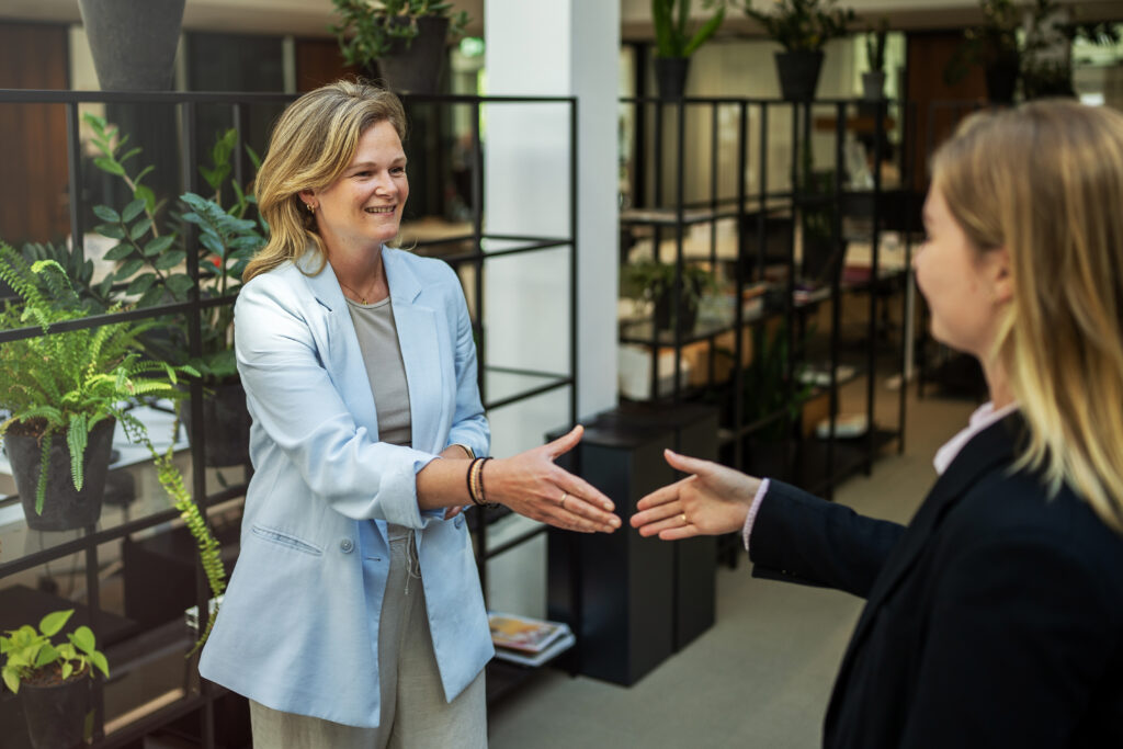 Twee vrouwen schudden handen in een modern kantoor met veel groene planten, terwijl ze elkaar vriendelijk begroeten. De vrouw links draagt een lichtblauwe blazer en glimlacht naar haar collega.