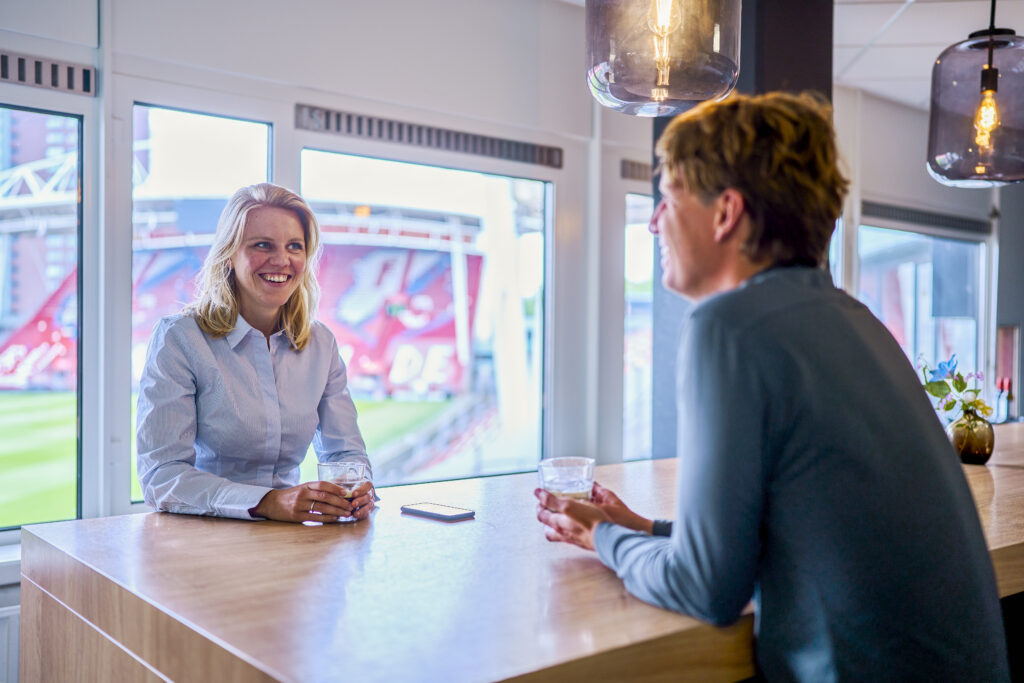 Twee collega's zitten aan de bar op kantoor, in een lichte ruimte bij het raam. Ze zijn lachend met elkaar in gesprek.