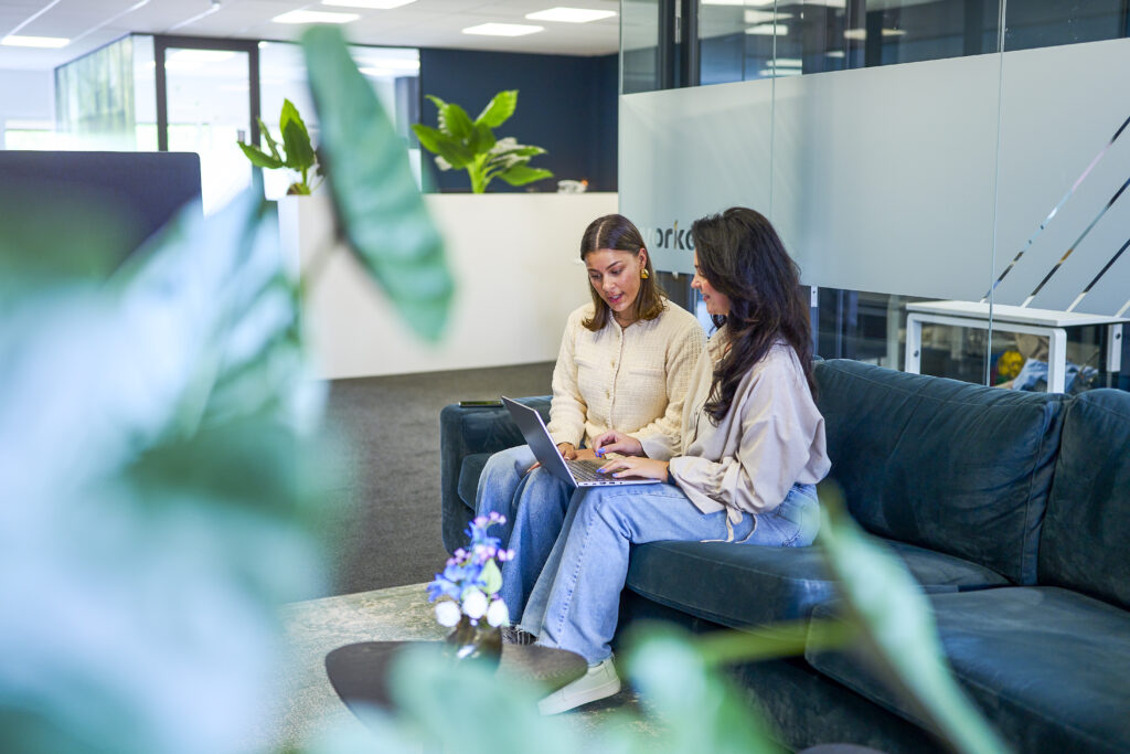 Twee vrouwen zitten op een groene bank in een moderne kantoorruimte en kijken samen naar een laptop. De vrouw links draagt een lichte trui en de vrouw rechts draagt een lichte blouse. Ze zijn verdiept in hun werk aan finance vacatures, terwijl de omgeving is gevuld met planten voor een frisse uitstraling.
